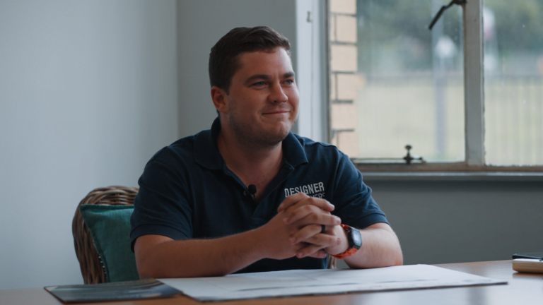 A man sitting at a desk, smiling slightly, with his hands clasped in front of him; papers and a window behind him. he wears a shirt labeled "designer.