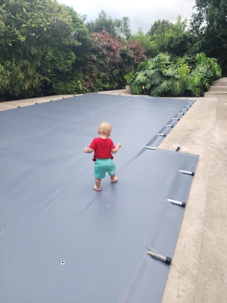 A toddler in a red shirt and green shorts walks on a large, covered surface outdoors, surrounded by dense green foliage, enjoying the ease of use of the child-friendly design that prioritizes pool safety.