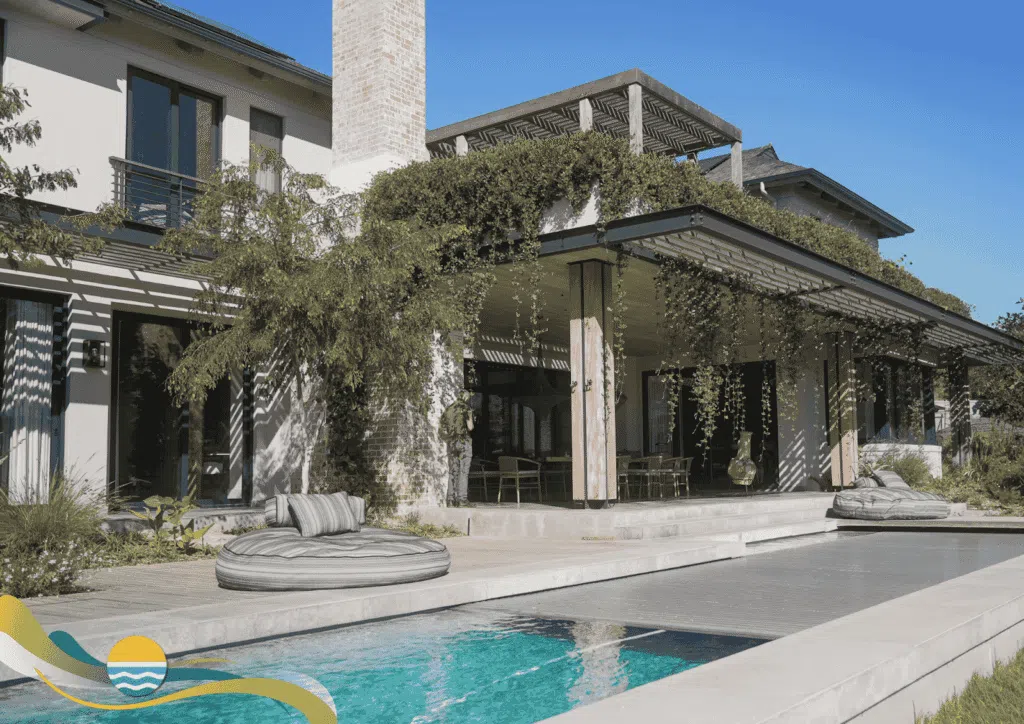 Modern house with greenery-covered pergola, outdoor seating area, and a swimming pool in the foreground under a clear blue sky.